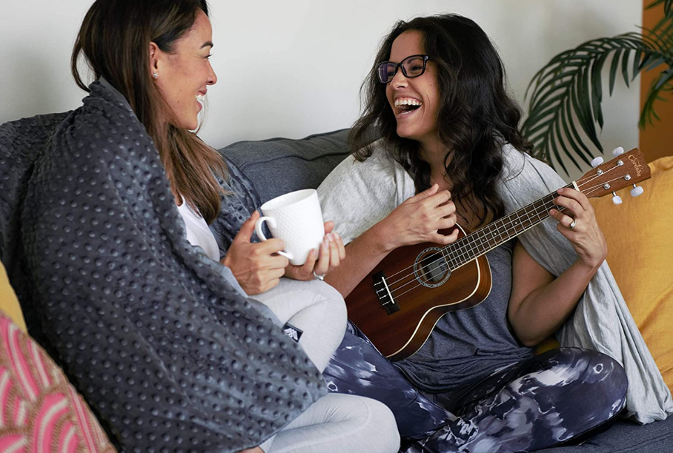 2 women sitting on couch under blanket laughing with one playing guitar