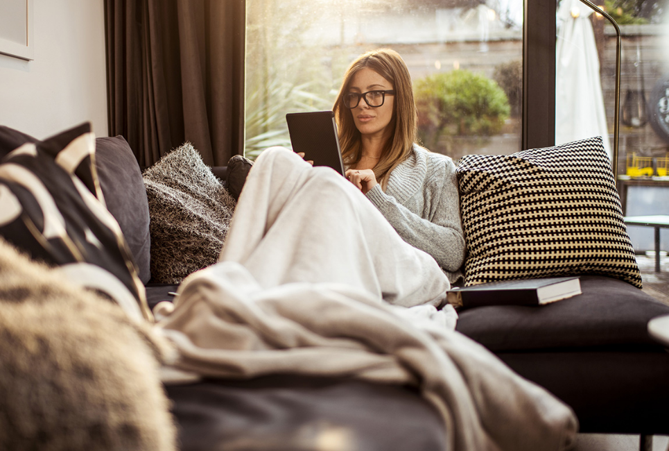 woman resting on couch reading on tablet with blanket on her legs