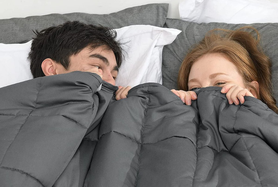 couple giggling in bed with grey weighted blanket pulled up to their faces