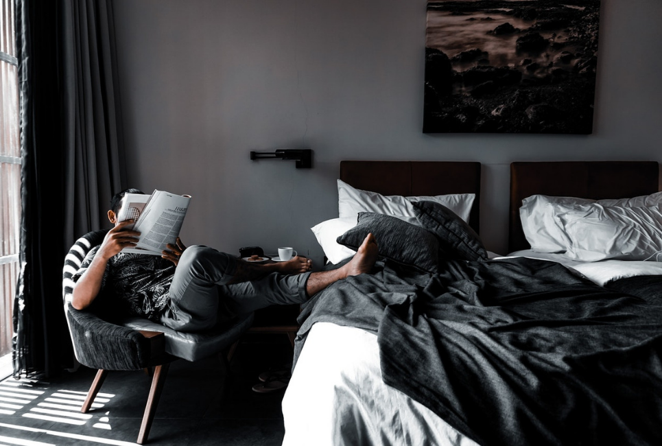man sitting in chair reading next to unmade bed