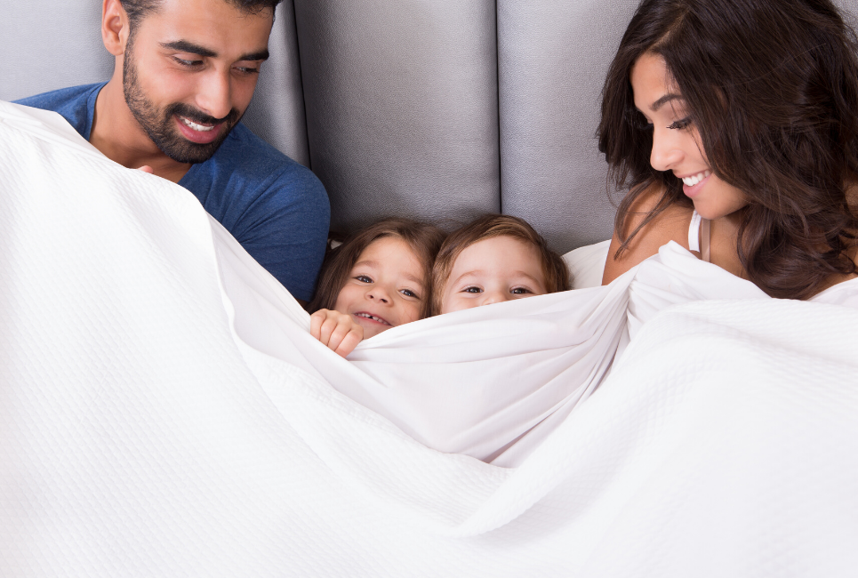 young family sitting up in bed together under blanket