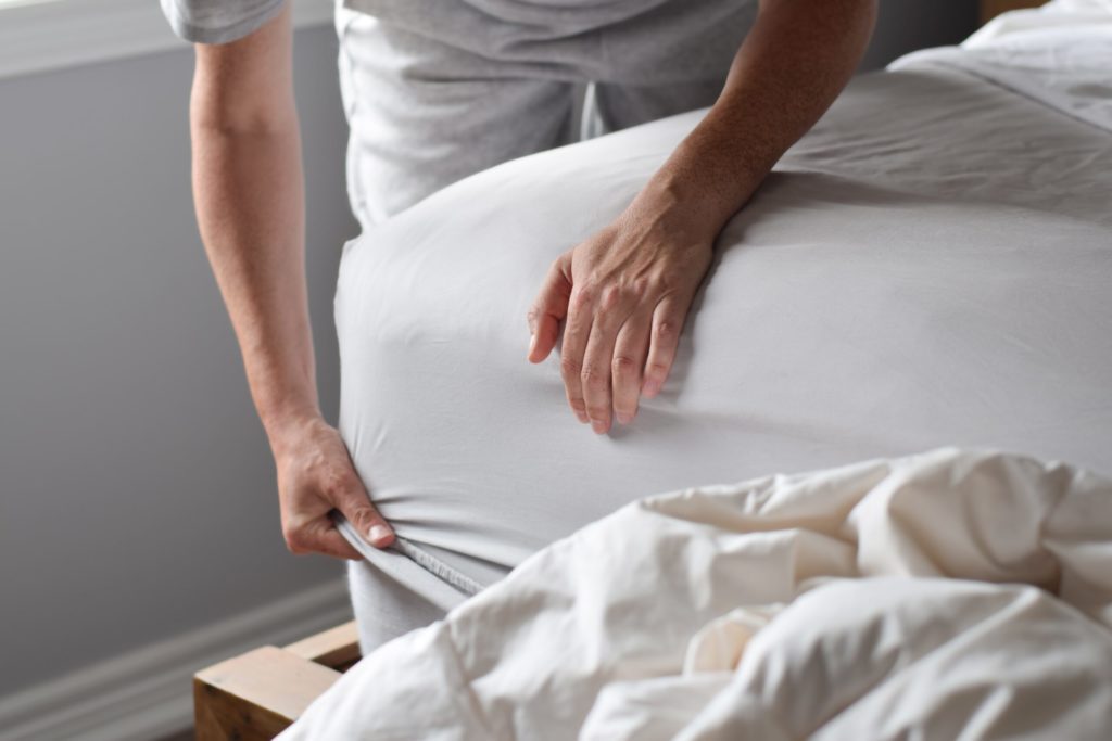 persons hand putting white sheets on corner of bed